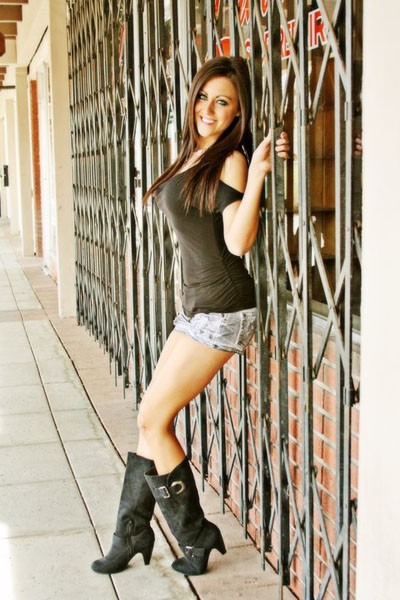 Hallie M is a model leaning against a security gate wearing cowboy boots, short shorts and a sleeveless knit top.